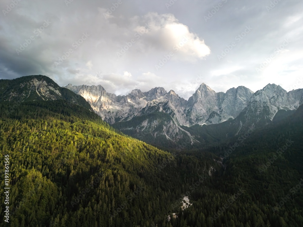 Obraz premium a valley with green forests and open fields, surrounded by mountain ranges. Sunlight breaks through the clouds, creating a soft contrast between light and shadow. Application: tourism, nature, ecology