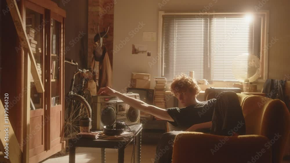 Melancholic young man lounging on sofa in warmly lit room, inserting audio cassette and adjusting volume on vintage boombox, listening to music to solace from his thoughts at home. Zoom shot