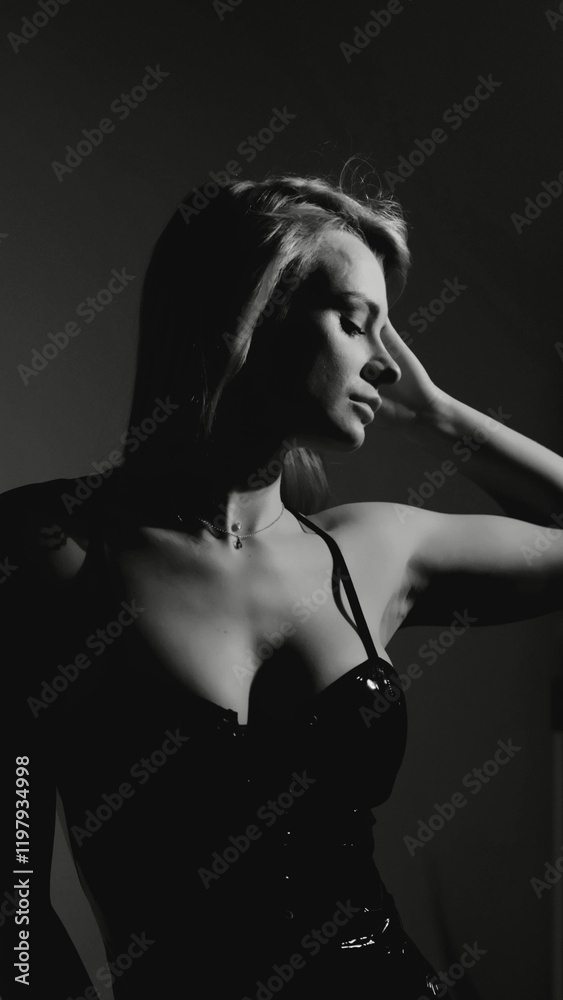 Fototapeta premium Blonde with a beautiful figure in a black patent leather bodysuit posing in a studio. Studio lighting, low exposure, monochrome image