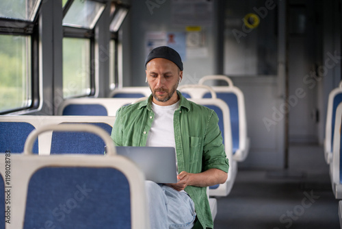 Thoughtful man passenger with laptop during electric train journey concentrating on tasks. Focused male freelancer in earphones remote works, productive use of travel time by railway public transport photo