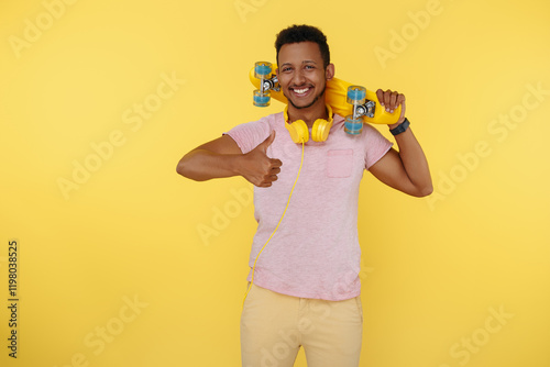 Careless african american young man wearing headphones holding skateboard over yellow background. Show thumb up. photo