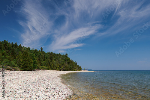 Along a stone beach, the blue and turquoise waters of a rocky bay out to a cape point, surrounded by lush green forests under a gorgeous blue sky with wispy clouds. 