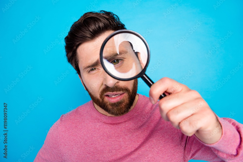 Curious bearded man examining through magnifying glass over blue background in casual wear Curious bearded man examining through magnifying glass over blue background in casual wear