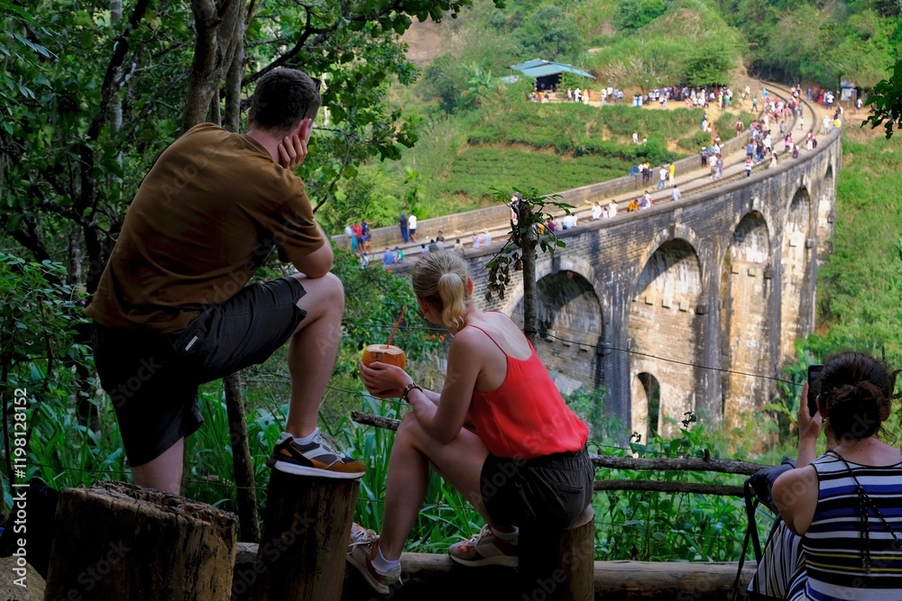 Silhouettes of young woman and man with coconut in hands sitting on viewpoint with iconic Nine Arch Bridge in Ella, Nuwara Eliya, Sri Lanka