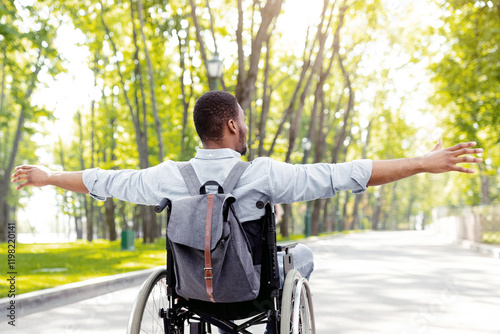 Back view of young disabled black man in wheelchair spreading his arms, feeling happy and free on walk at city park. Full length of handicapped guy with backpack going on trip outdoors