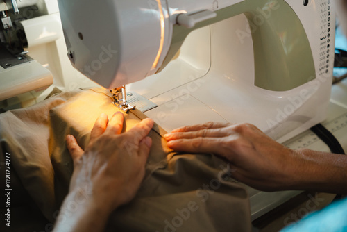 Close-up of woman hands sewing fabric on a sewing machine. The fabric is a light brown color, and the machine is white.