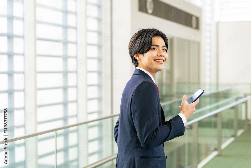 スマホ、ビジネスマン、スマートフォン、男性、会社員、オフィス、アプリ、SNS、メール、検索、モバイル、動画、笑顔、ゲーム、ソーシャル、ネットワーク、通信、回線、電波、wi-fi、無線、ワイヤレス、操作、使う、見る、ロビー、暇つぶし、社内、LAN、ビル、屋内、日本人、ビジネス、DX、デジタル、インターネット、オンライン、web、予約、情報、ニュース、携帯電話、サラリーマン、スーツ、待ち合わせ、駅、空