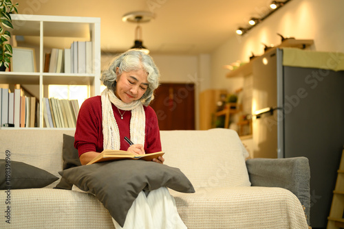 Calm senior woman in a red sweater sitting on couch and writing in journal photo