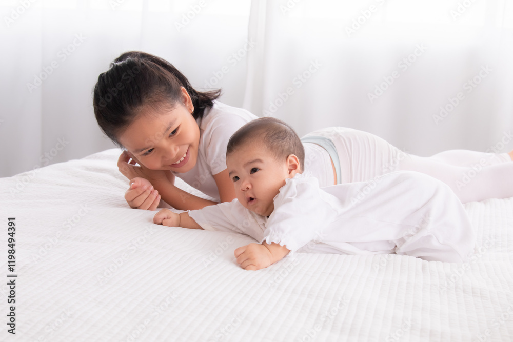Asian smiling young girl lying next to baby on white bed in bright indoor setting. Both wearing white clothing, sharing a joyful bonding moment in a calm and cozy home environment. Asian smiling young girl lying next to baby on white bed in bright indoor setting. Both wearing white clothing, sharing a joyful bonding moment in a calm and cozy home environment.