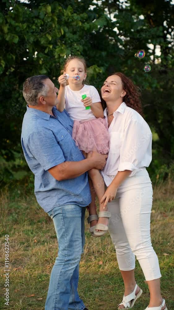Middle aged mom and dad hold their daughter in their arms, she is having fun in park, blowing soap bubbles. Outdoor games, summer holidays, weekends. International Day of Families Middle aged mom and dad hold their daughter in their arms, she is having fun in park, blowing soap bubbles. Outdoor games, summer holidays, weekends. International Day of Families