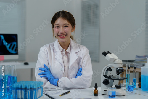 Young scientist wearing lab coat and gloves smiles with crossed arms in modern laboratory with microscope, test tubes, and computer displaying dna double helix structure photo