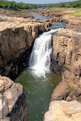 A picturesque waterfall cascading over rocky cliffs into a serene pool, surrounded by lush greenery under a clear blue sky.