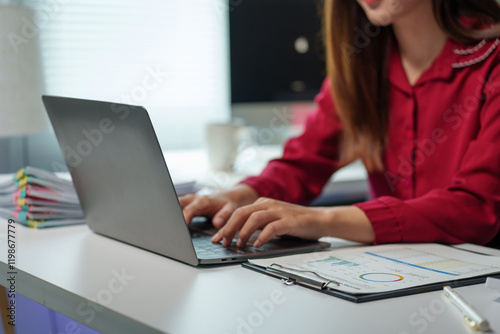 Businesswoman dressed in a red shirt is typing on a laptop keyboard while analyzing financial reports and charts in a sleek, modern office environment