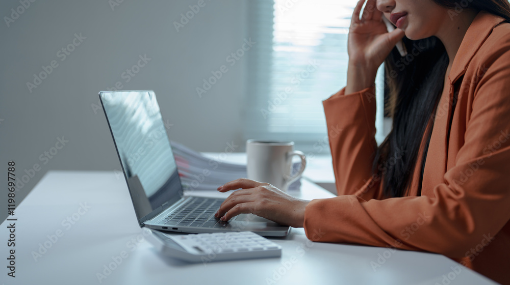 Asian businesswoman wearing an orange blazer is working on a laptop and talking on the phone in a modern office, with a calculator and documents on the desk Asian businesswoman wearing an orange blazer is working on a laptop and talking on the phone in a modern office, with a calculator and documents on the desk