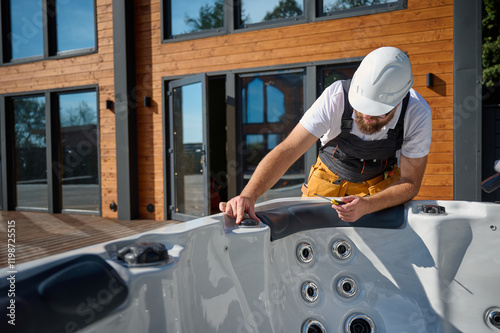 Plumber installing hot tub. Spa technician performing hot tub. Bathtub plumbing. Plumber fixing spa zone. Professional in uniform inspects and prepares a hot tub located on a stylish outdoor patio.