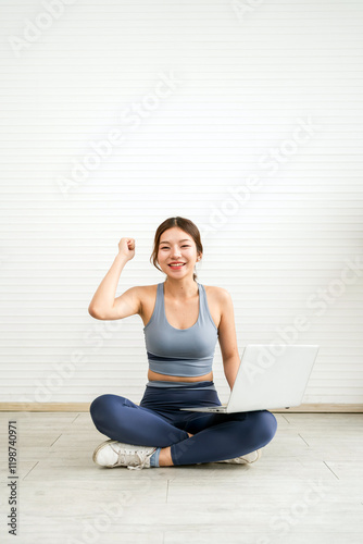A woman in active wear sits on the floor, smiling and raising her hand, with a laptop in front of her, conveying a sense of success and motivation. Raised fist and a bright smile.