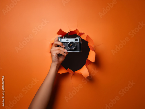 A hand holds a vintage camera through a torn hole in vibrant orange paper. The contrast creates a striking visual. photo