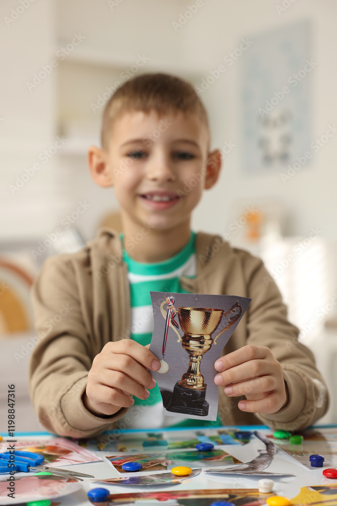 Creating vision board. Little boy with picture of golden cup trophy at table indoors Creating vision board. Little boy with picture of golden cup trophy at table indoors