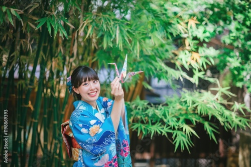 A young woman in a vibrant blue kimono holds a delicate origami crane amidst lush bamboo. photo