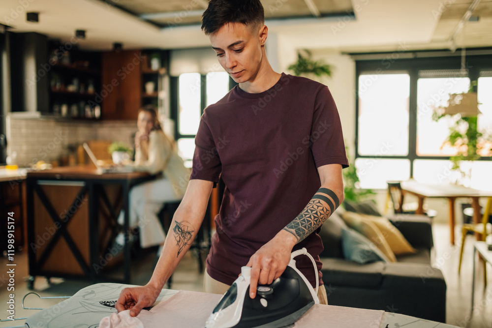 Obraz premium Young woman ironing clothes at home while her partner works on laptop in the background