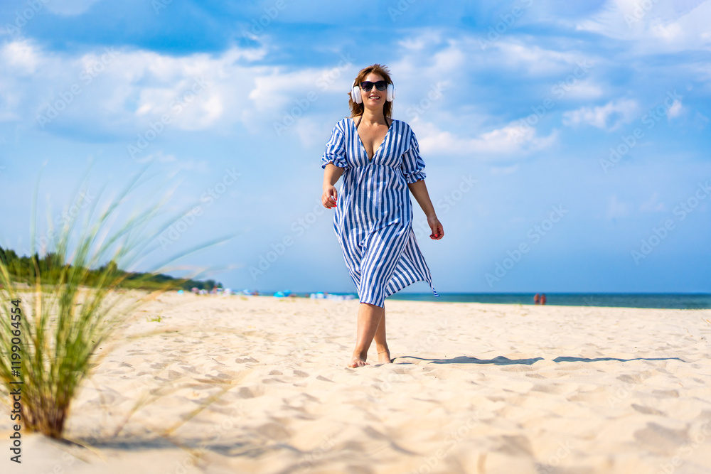 Vacations with music. Beautiful mature woman walking on sandy beach and listening to music in headphones in summer Vacations with music. Beautiful mature woman walking on sandy beach and listening to music in headphones in summer