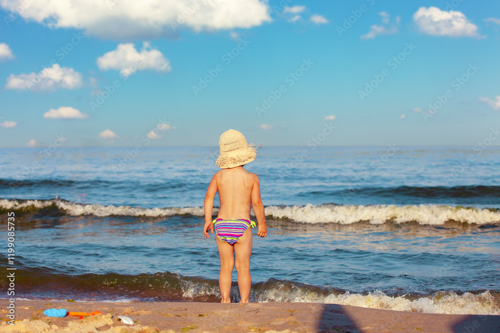 Happy little girl standing on the beach in summer with her back to the camera. The girl is looking at the sea. The baby in a straw hat