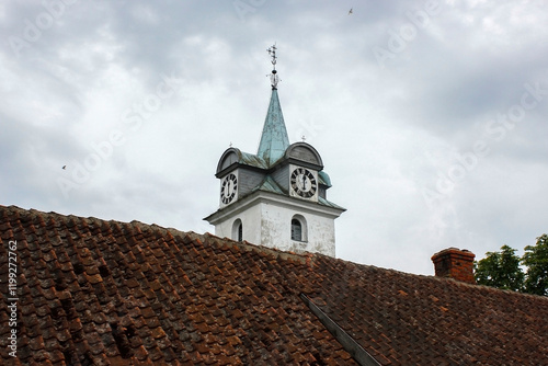 A white church tower in a small European city from the side with a visible clock and a metal roof with a Christian cross on top of it. The clock shows 6 o'clock time.