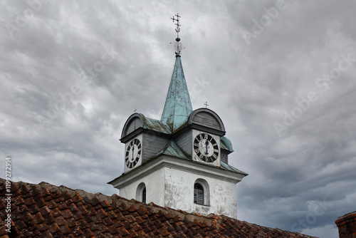 A white church tower in a small European city from the side with a visible clock and a metal roof with a Christian cross on top of it. The clock shows 6 o'clock time.