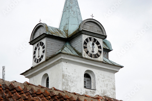 A white church tower in a small European city from the side with a visible clock and a metal roof with a Christian cross on top of it. The clock shows 6 o'clock time.