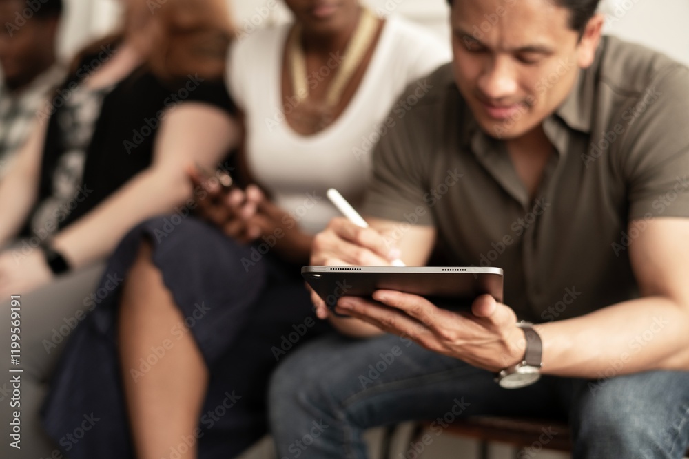 A diverse group of people sitting together. Man uses a tablet, focusing. The group, including a woman, is engaged with technology. Diverse, engaged, technology-focused. Latin man using social media. A diverse group of people sitting together. Man uses a tablet, focusing. The group, including a woman, is engaged with technology. Diverse, engaged, technology-focused. Latin man using social media.