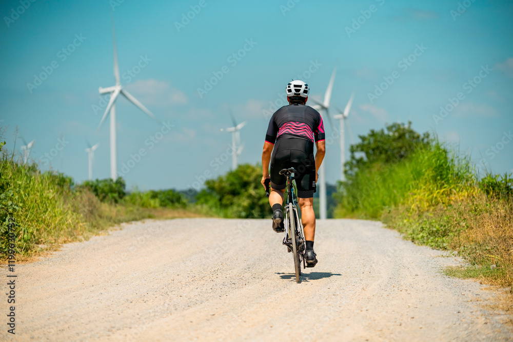 Fototapeta premium Cyclist riding on a dirt road surrounded by wind turbines on a sunny day