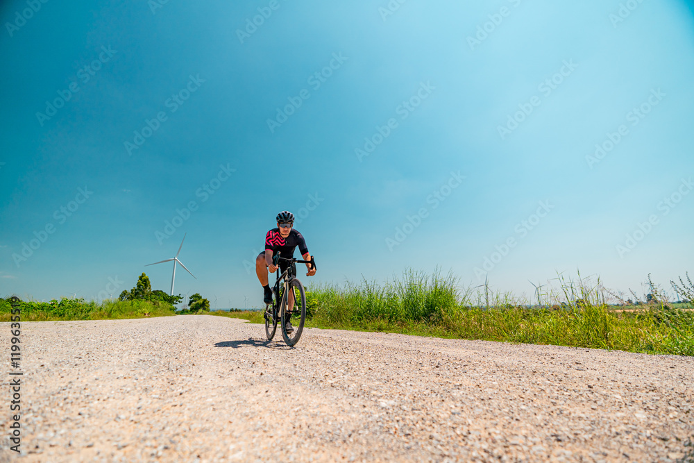 Fototapeta premium Cyclist rides on a gravel road surrounded by green fields and wind turbines under a clear blue sky
