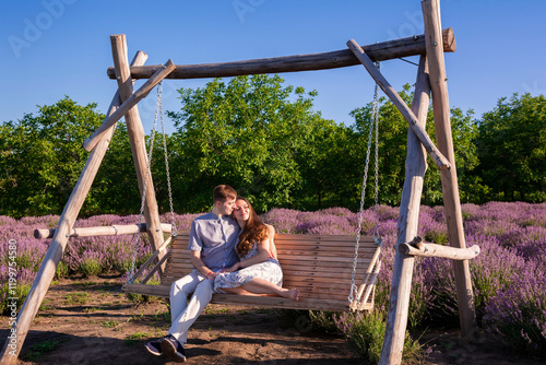 Lovely couple sits on a rustic wooden swing amidst lavender field during a sunny summer day