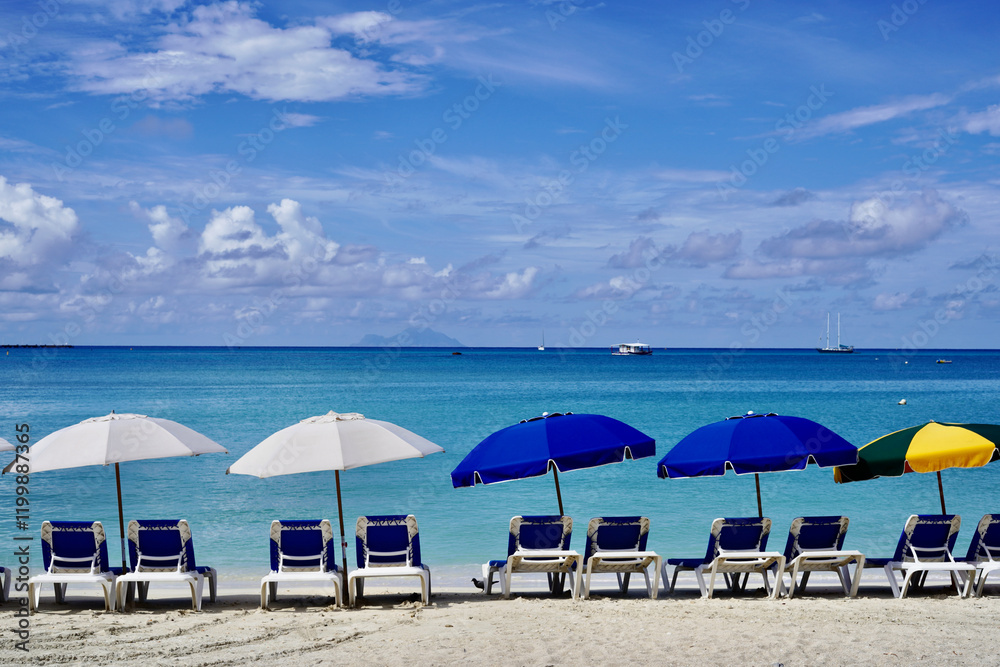 Beach loungers along Great Bay Beach await sunbathers at the start of a sunny day in St. Maarten against a backdrop of calm turquoise water turning to deep blue at the horizon.