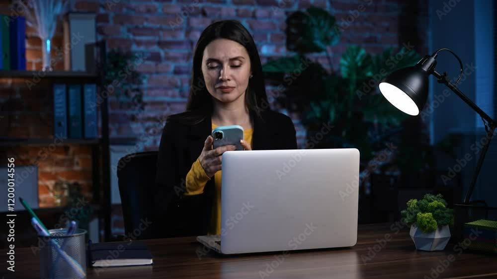 Young business woman sitting at desk working late in office loft, using laptop, texting sending messages on smartphone, chatting smiling expression.