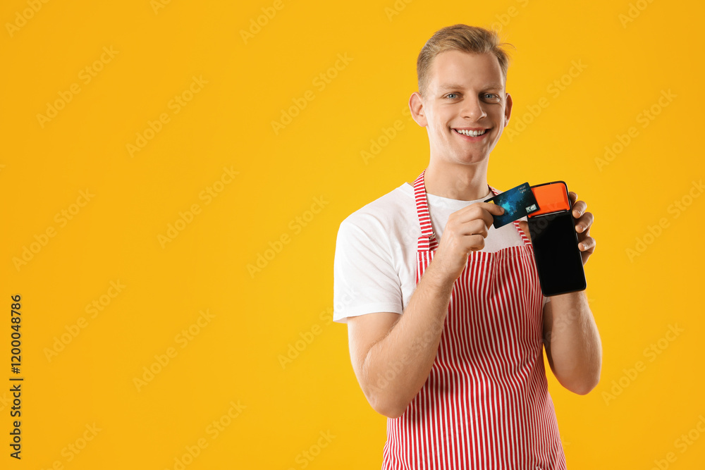 Happy young man in apron with payment terminal and debit card on yellow background, space for text Happy young man in apron with payment terminal and debit card on yellow background, space for text