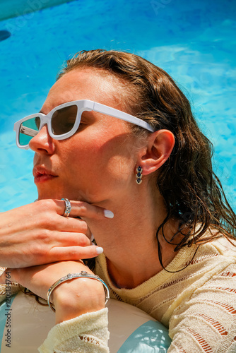 A young girl swims in a swimming pool on a striped blue and white inflatable circle. A girl in blue sunglasses sunbathes in the sun.
