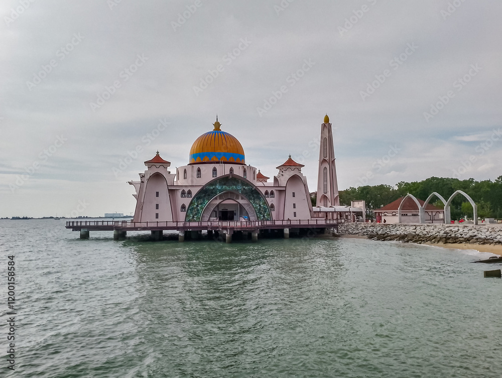 Fototapeta premium Scenic view of iconic white Malacca Straits Mosque stands partially in water against vibrant sky in Malaysia, Southeast Asia. Golden dome and tall minaret are prominent. Religious muslim landmark
