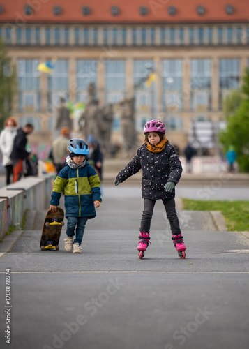 Girl is skating on roller skates, little boy is walking next to her with skateboard in his hands. Children having fun outdoors in cold season. photo