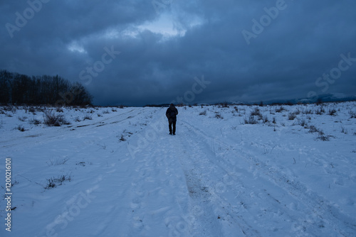 Tourist snow road. An unrecognizable man is walking along the road with a backpack. Dark blue saturated clouds. A winter atmospheric mood for walking in the countryside. Active lifestyle, walking