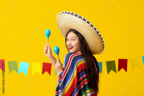 Happy young woman in Mexican sombrero hat and poncho holding maracas on yellow background