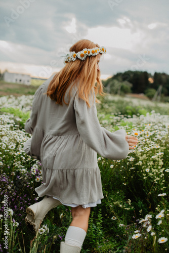A young woman in a flowy dress and headband walks through a blooming daisy field, enjoying the peaceful countryside atmosphere under a cloudy sky.

