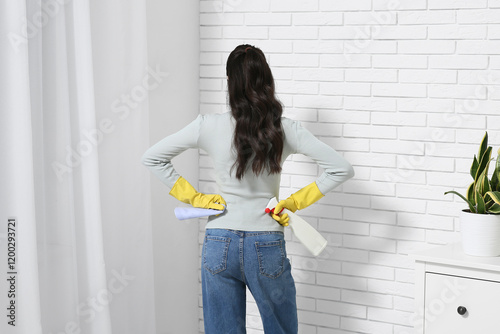 Young woman with cleaning supplies looking at mold on white brick wall, back view photo