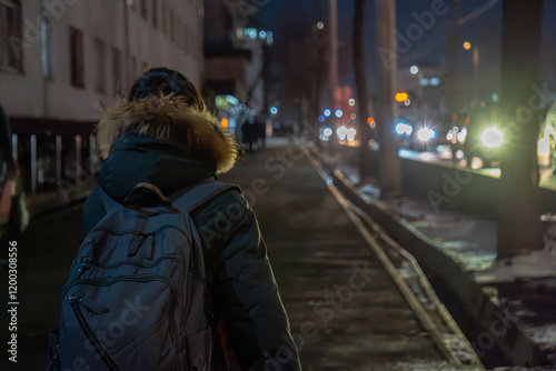 A person walking on a dimly lit urban sidewalk at night, carrying a backpack and wearing a fur-lined jacket. Car headlights and streetlights illuminate the blurred background, creating a moody scene