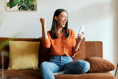 Euphoric lady getting good news notification via smartphone, making yes gesture and shouting, celebrating success while sitting on couch at home