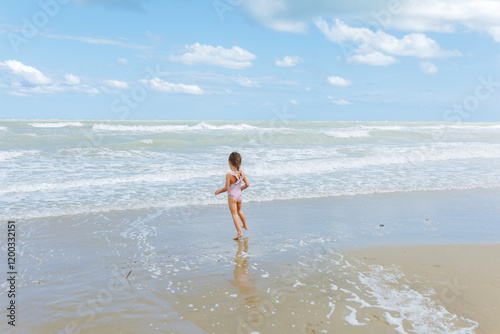 rea view of little girl in the swimsuit jumping on the beach playing with waves