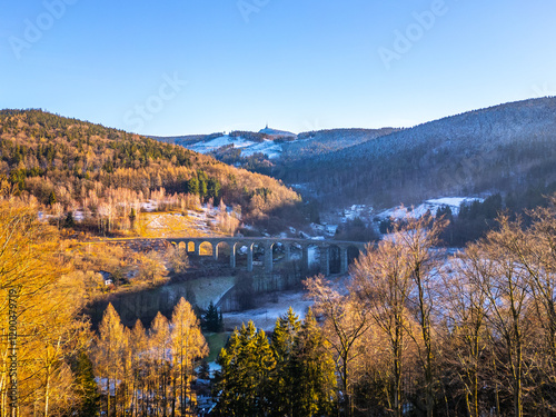 A picturesque railway stone bridge stands majestically in Novina, Krystofovo udoli, against the backdrop of Jested Mountain at winter sunset, illuminating the serene landscape with warm hues. photo