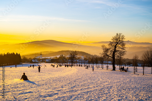 Sunset at Prichovice showcases vibrant colors over snow-covered hills. People sled and enjoy the winter landscape, creating a cheerful atmosphere in the crisp evening air. photo