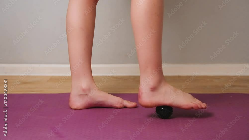 Teen girl kid feet are doing foot exercises by rolling a massage black ball under the foot on yoga mat on floor indoors home interior. Training to correct and relax the foot, posture. Close-up Teen girl kid feet are doing foot exercises by rolling a massage black ball under the foot on yoga mat on floor indoors home interior. Training to correct and relax the foot, posture. Close-up