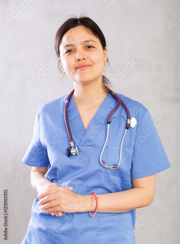 Confident positive young female doctor in blue uniform with stethoscope around neck standing indoors on gray background. Professional activity concept